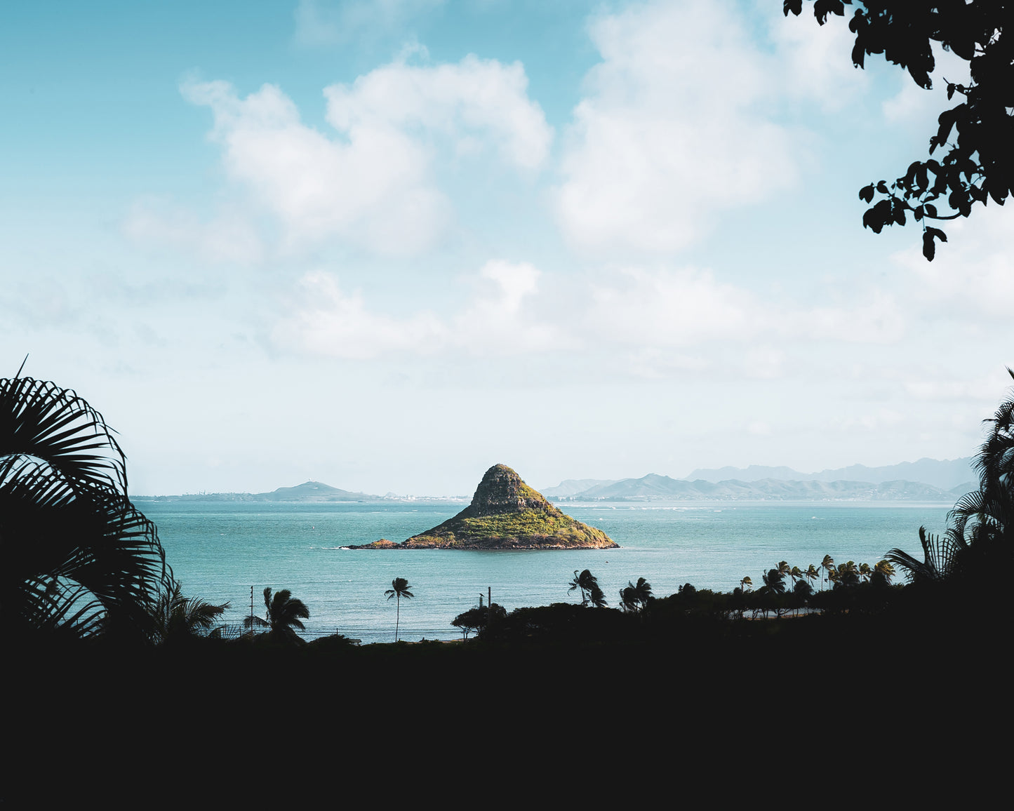A very green Chinaman's Hat (Mokoli'i) in Oahu, Hawaii, surrounded by clear turquoise ocean and framed with silhouetted palm trees and mountains in the distance.