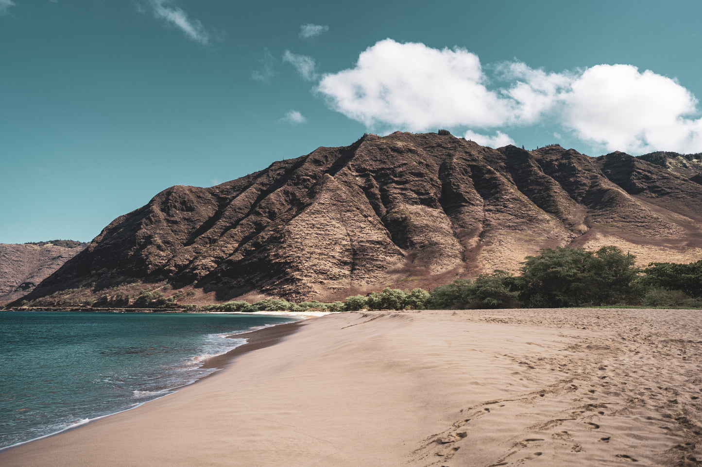 A pristine stretch of sand at Makua beach on the west side of Oahu, Hawaii, with clear blue waters meeting rocky mountains under a bright sky with scattered clouds.