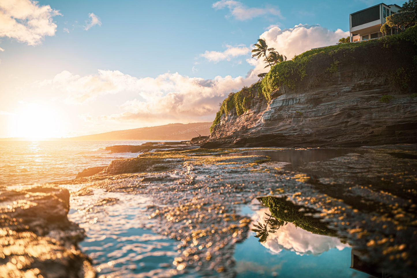 Golden sunset at Spitting Caves, Hawaii, with rugged cliffs, crashing waves, and a calm tide pool reflecting palm trees.