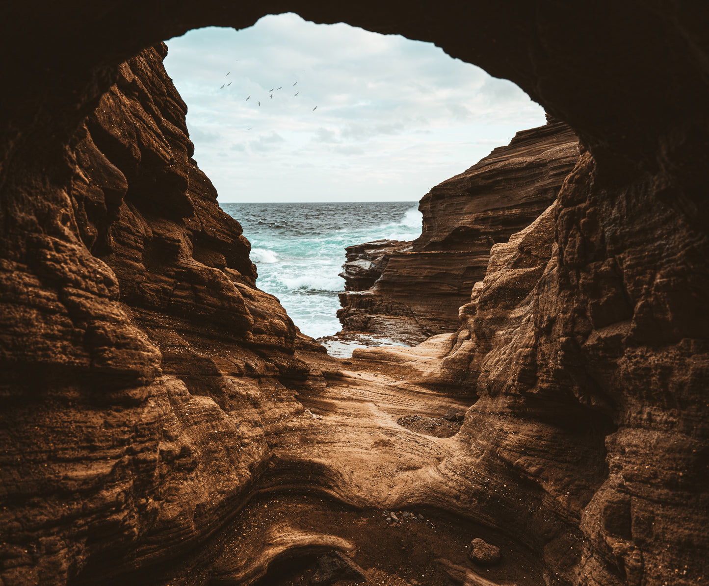 Inside of a lava tube looking out at rugged cliffs and crashing waves in Oahu, Hawaii.