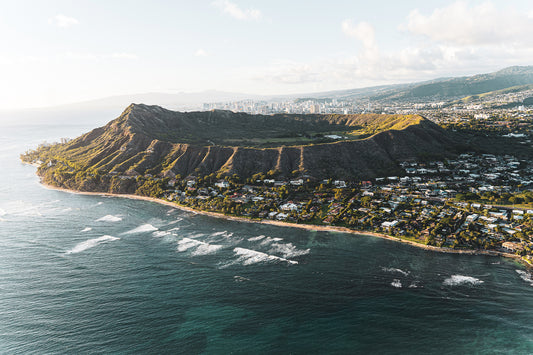 Aerial view of Diamond Head Crater on the island of Oahu, Hawaii, with the coastline and ocean in the foreground. The crater contrasts with the surrounding neighborhoods and city skyline of Honolulu in the background.