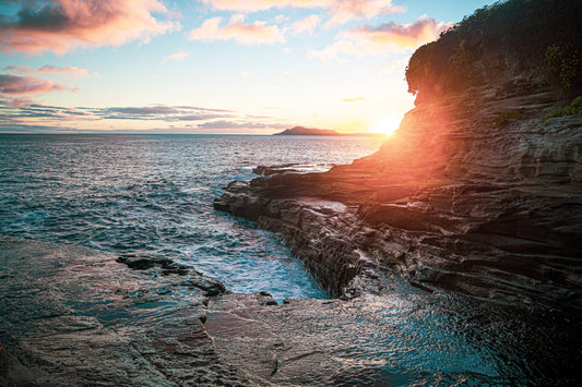 Sunset at Spitting Caves in Oahu, Hawaii, with waves hitting layered cliffs and golden light illuminating the coastline.