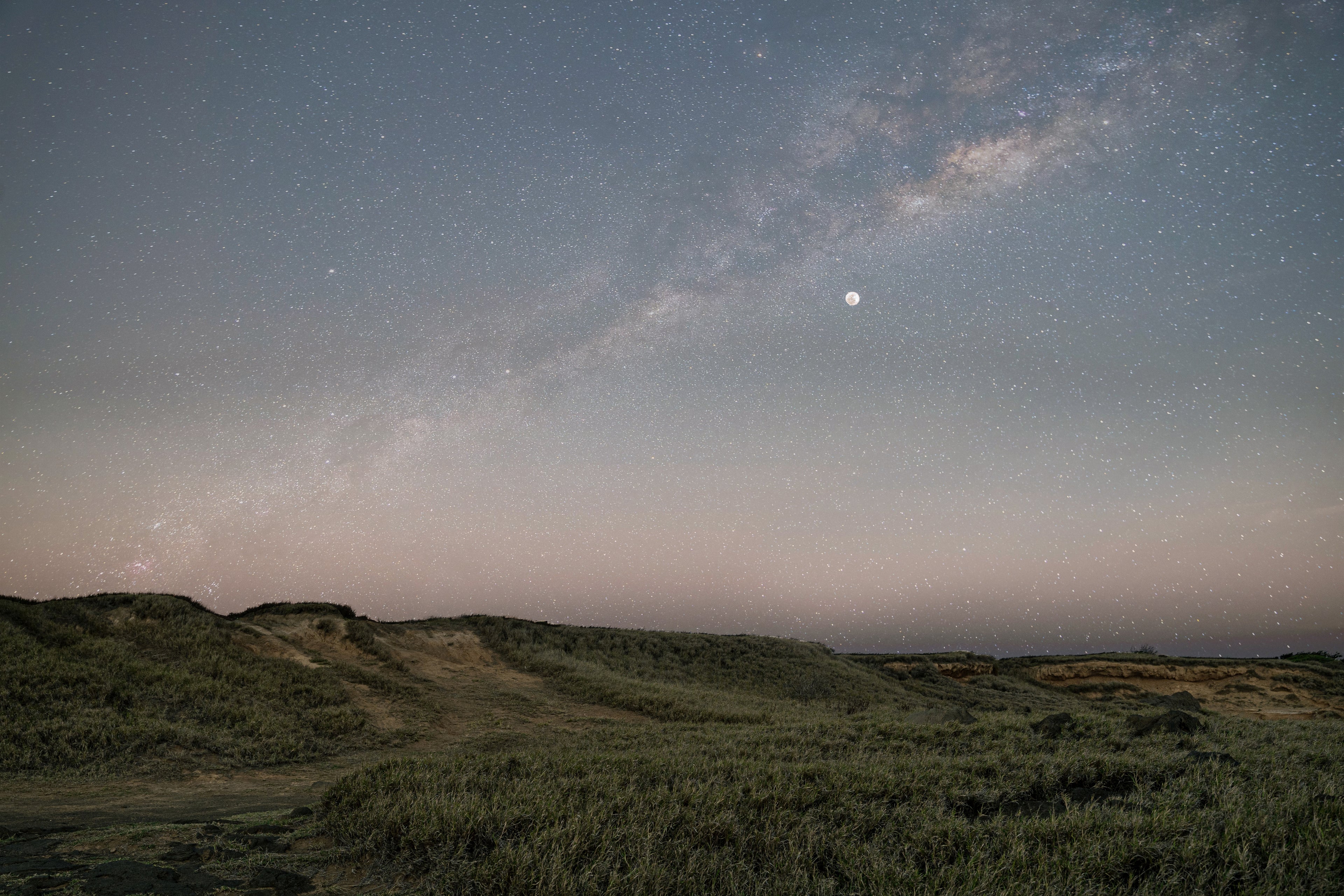 A serene night sky filled with twinkling stars and a bright moon next to the milky way illuminating a landscape of grassy sand dunes.