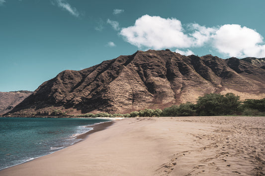 A pristine stretch of sand at Makua beach on the west side of Oahu, Hawaii, with clear blue waters meeting rocky mountains under a bright sky with scattered clouds.