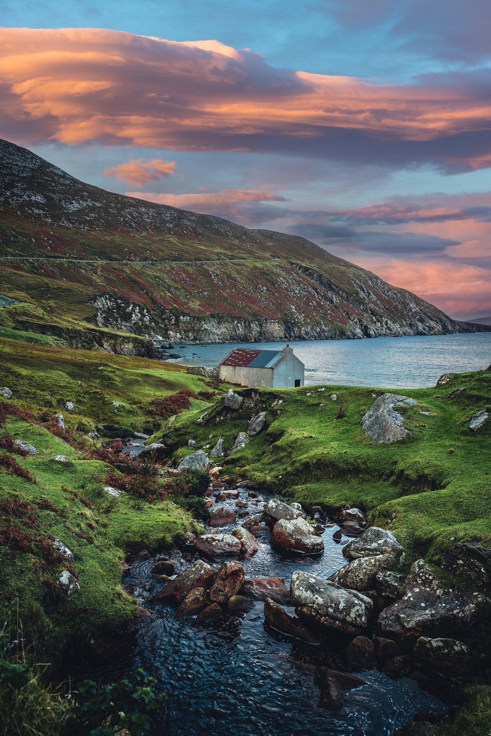 A small stone cottage by the sea at Keem Bay, Achill Island, Ireland, with a winding stream, green hills, and a vibrant pink-orange sunset.