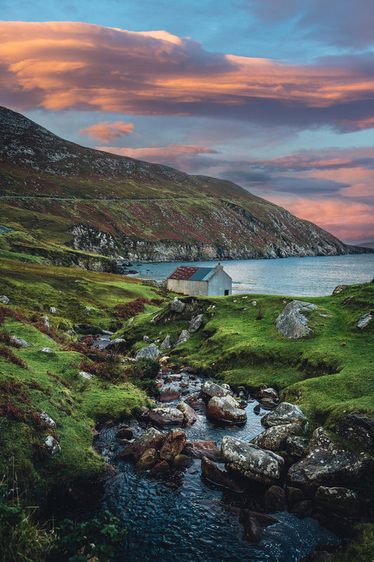 A small stone cottage by the sea at Keem Bay, Achill Island, Ireland, with a winding stream, green hills, and a vibrant pink-orange sunset.