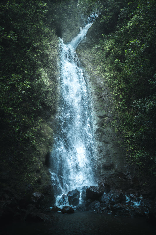Lulumahu Falls in Oahu, Hawaii, cascades down a lush, green mountain side into a rocky pool of water below.